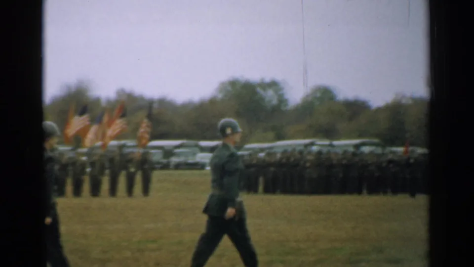 A uniformed man crosses the foreground as soldiers stand in formation with American flags behind him in the New Hampshire military scene.