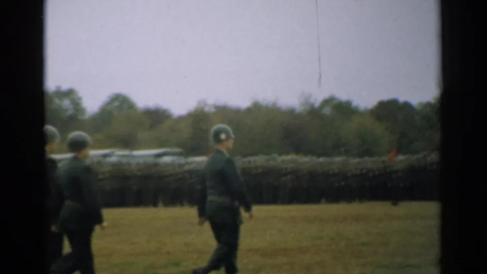 Two uniformed soldiers move across the foreground with a long military formation stretching behind them on the grassy parade field.