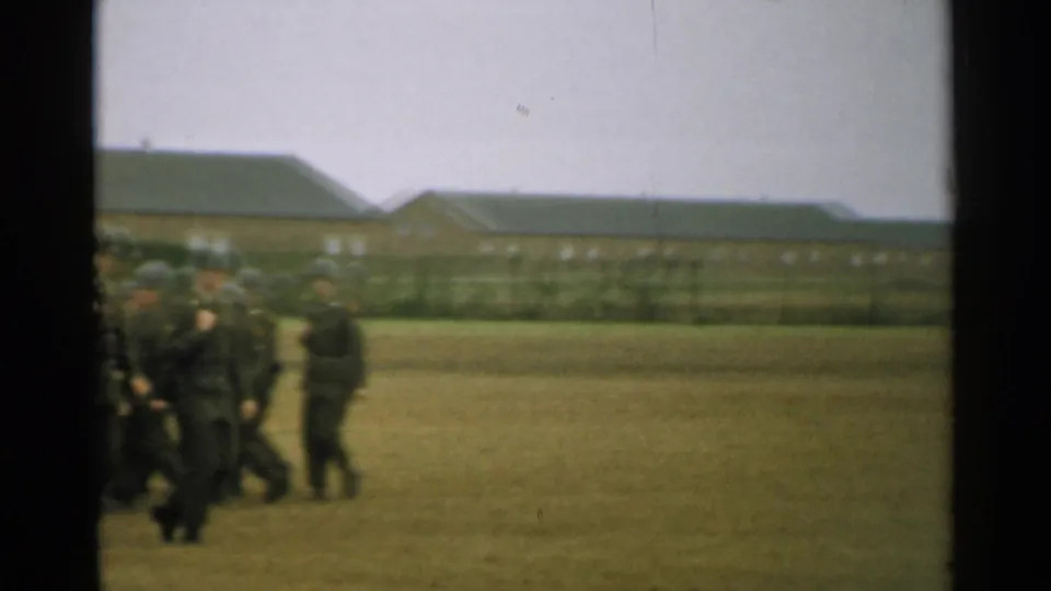 Soldiers in dark uniforms gather or march at the edge of the frame, with low institutional buildings beyond the parade field.