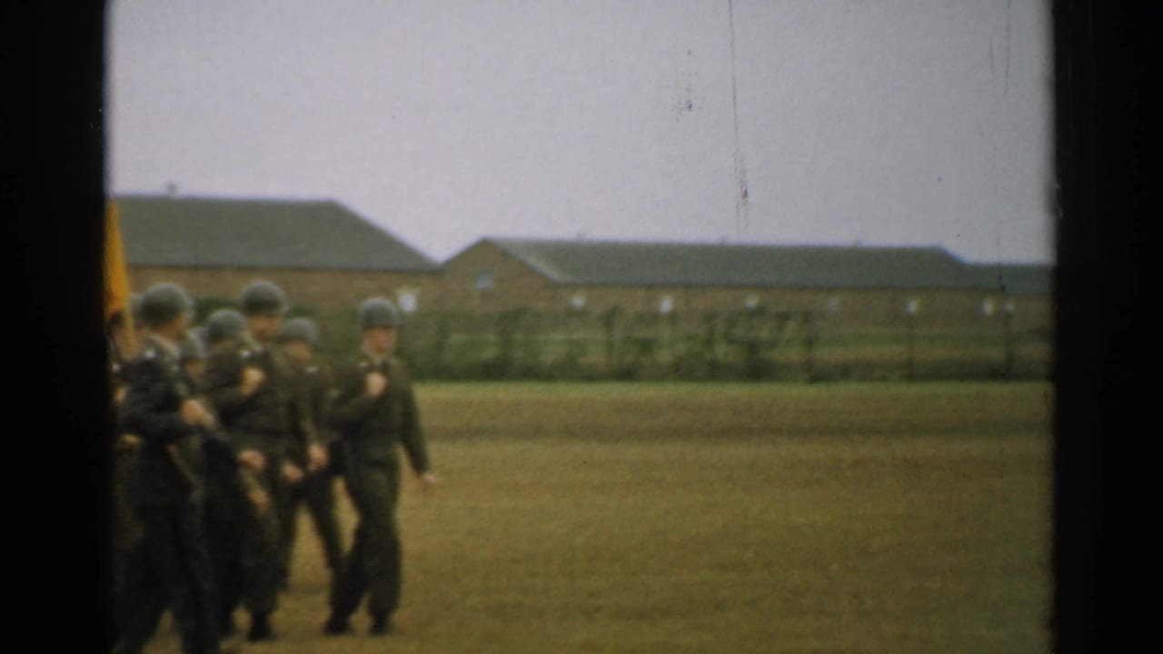 Soldiers in green helmets march across a grassy field near brick barracks.