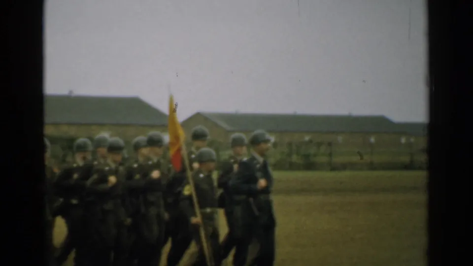 Helmeted soldiers in formation march across a New Hampshire military field in 1952, with a color flag near the front of the group.