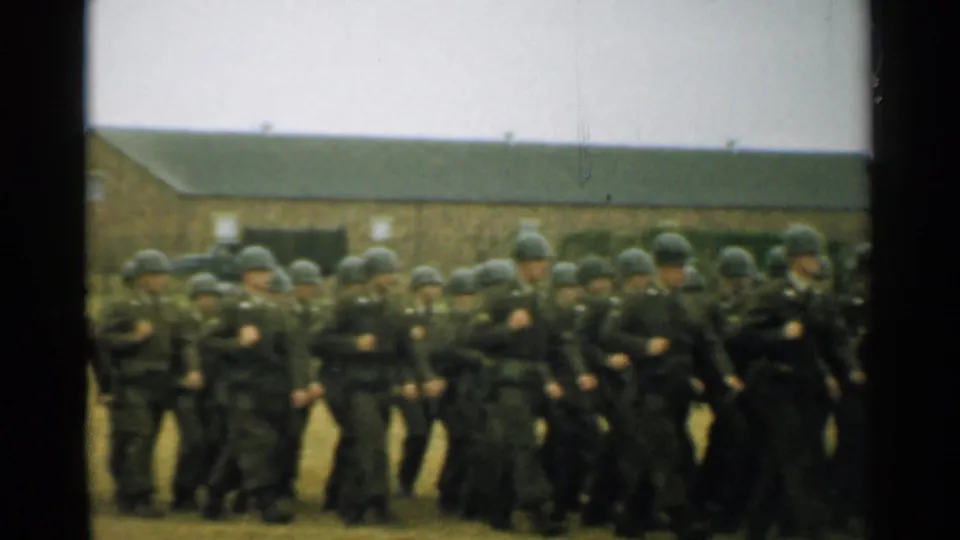 1952 New Hampshire frame showing a group of uniformed soldiers marching in step across a grassy base field before brick barracks.
