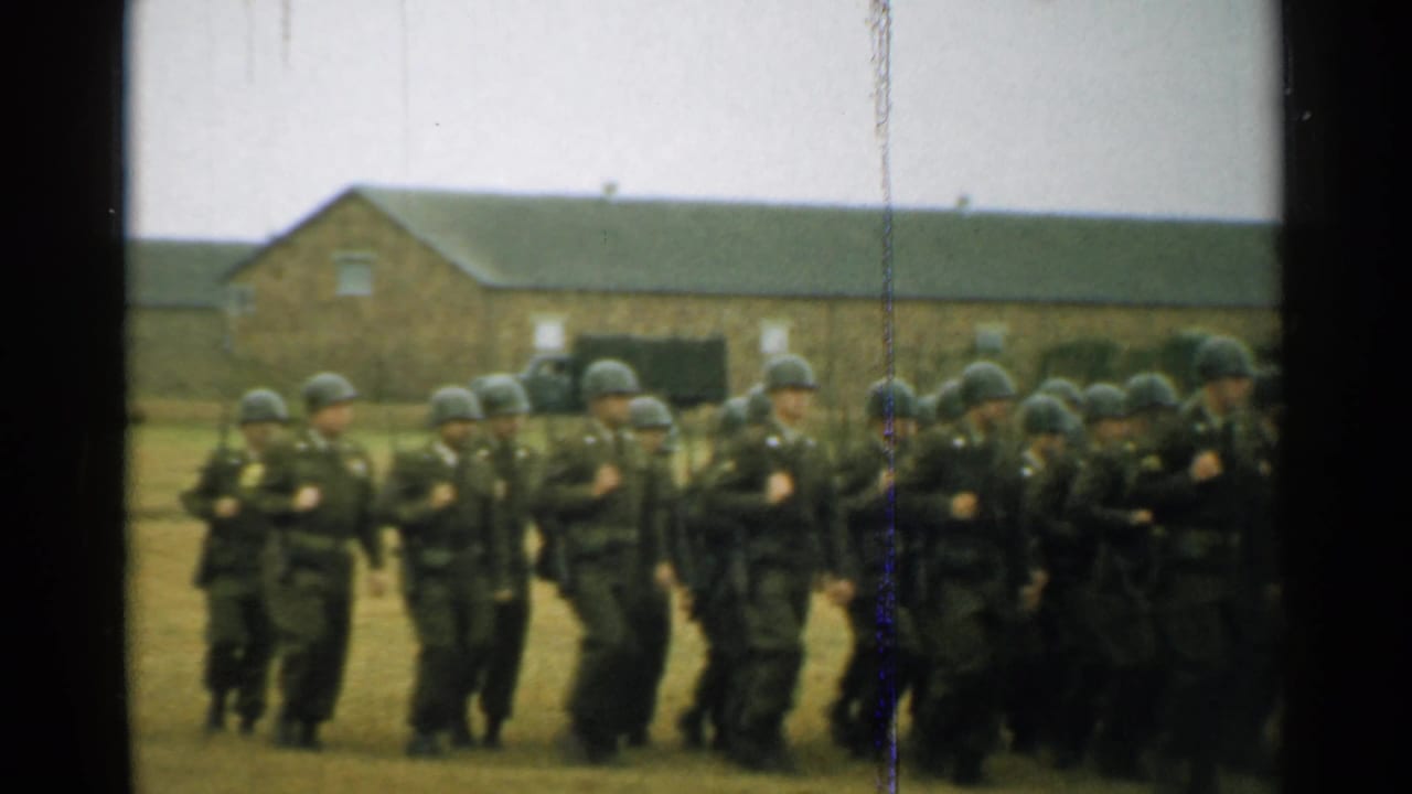 Helmeted soldiers march in formation across a grassy field near brick barracks.