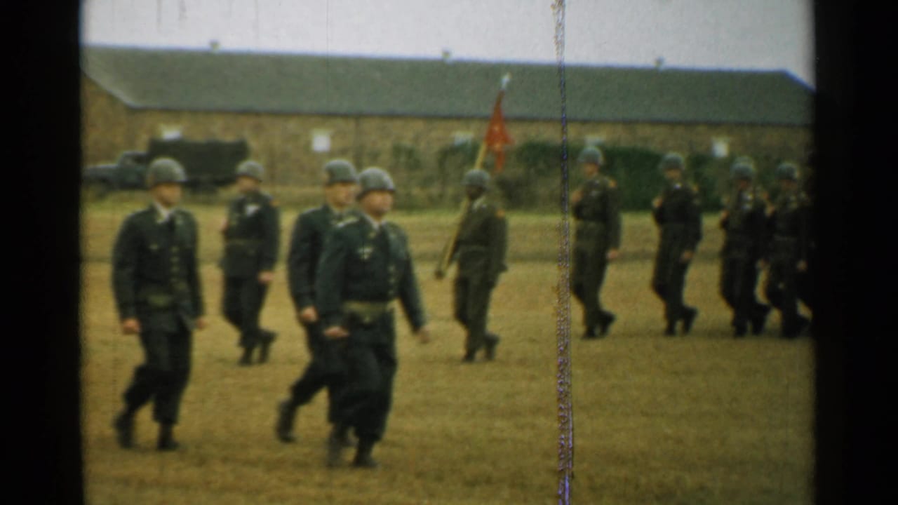 Soldiers in helmets march past with an orange flag visible behind them.