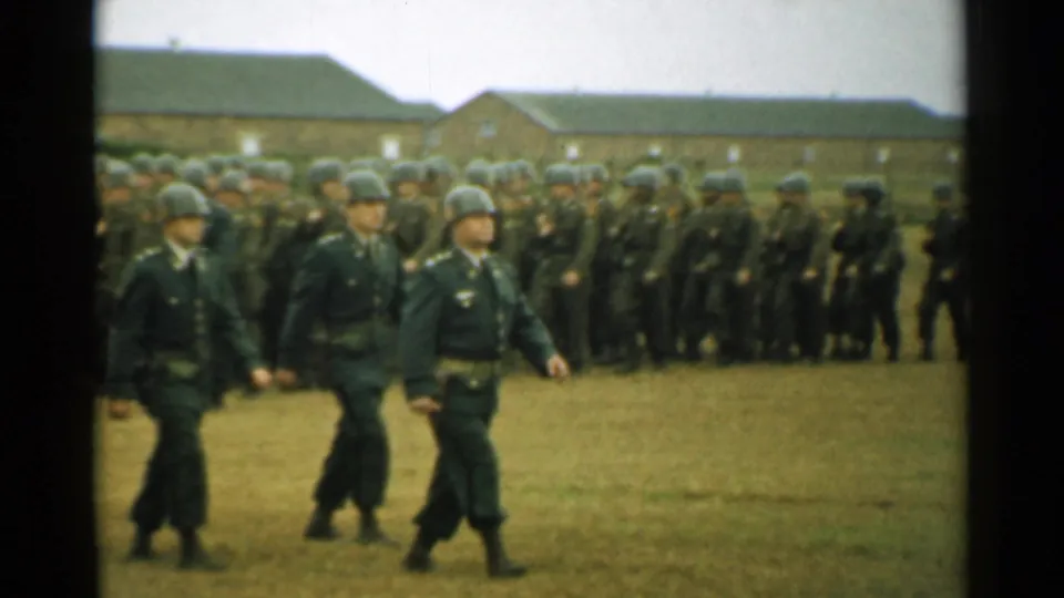 Helmeted soldiers march in the foreground with a larger formation and brick barracks behind them in New Hampshire 1952 footage.