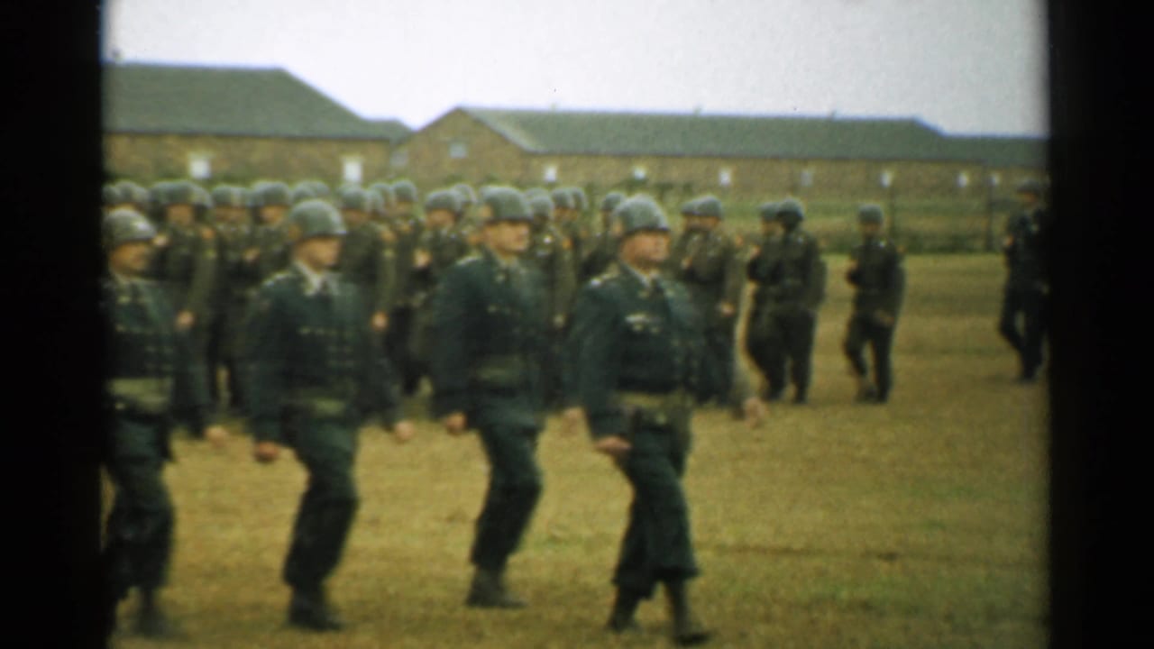 Soldiers in helmets march across a grassy field in front of brick barracks.