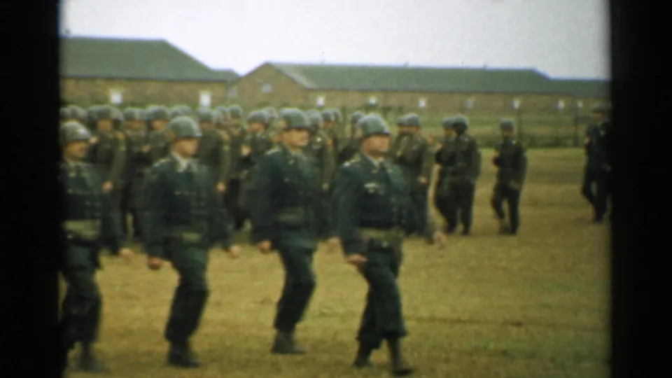 1952 New Hampshire home-movie frame showing uniformed soldiers in helmets marching past brick barracks on a grassy military field.