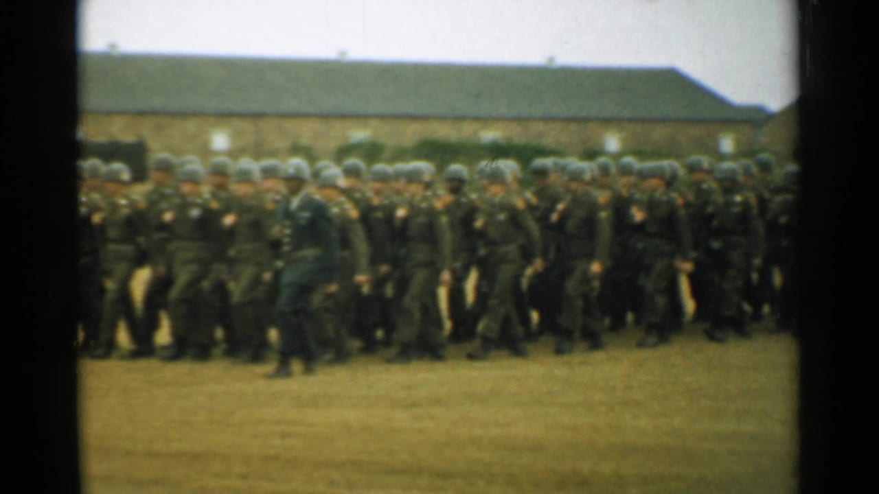 A large group of helmeted soldiers marches in rows on a field near barracks.