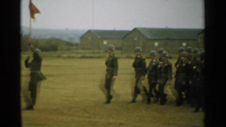 Soldiers in helmets pass a standing figure on a grassy military field, with a flag and brick barracks in the background.