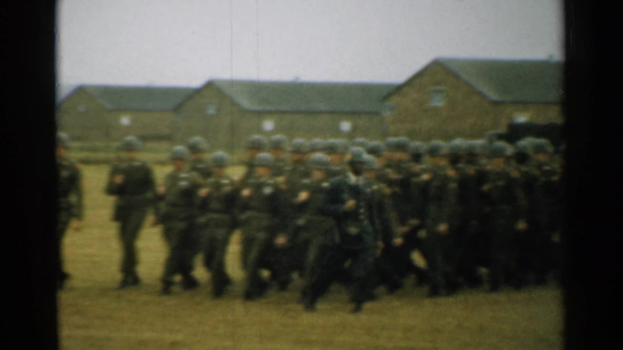 Soldiers in helmets march across a grassy field near brick barracks.