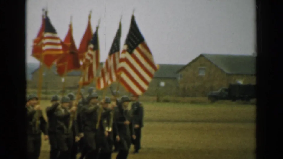 U.S. flags and red military banners fill the frame as soldiers march on a New Hampshire base in 1952.