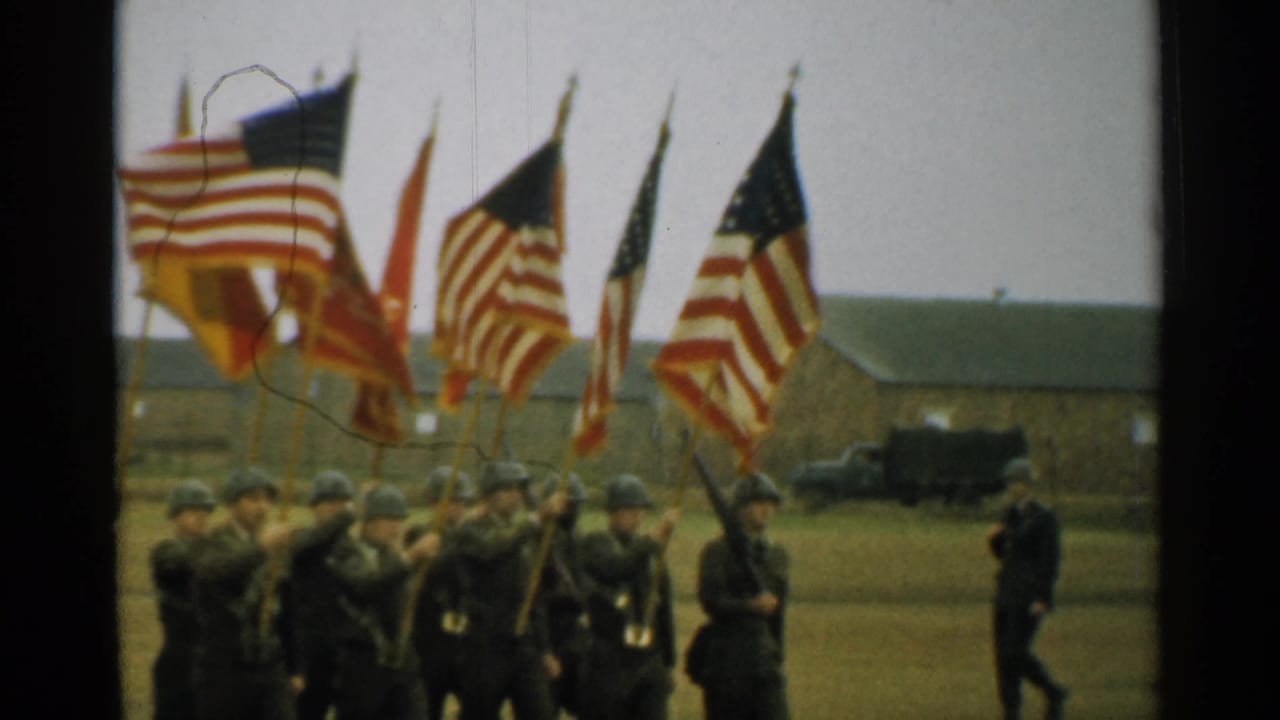Soldiers march across a field carrying American flags and military colors near barracks.