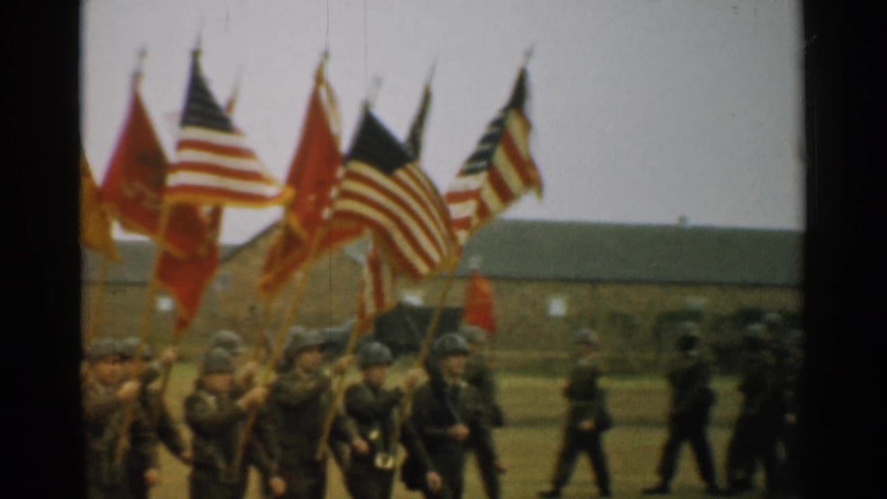 Army flag bearers march in formation with U.S. flags and red colors, with officers visible at the edge of the field.