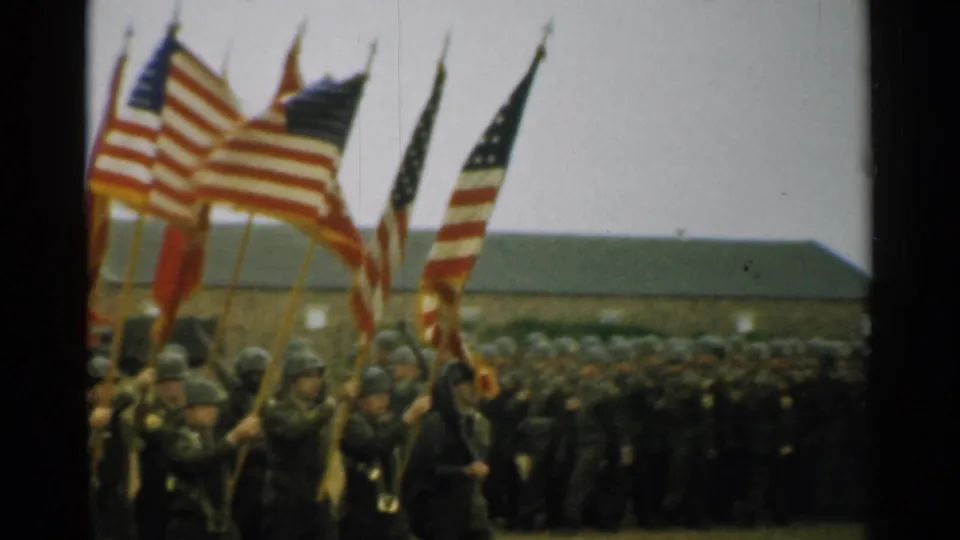 1952 home-movie frame of a dense Army formation following flag bearers across a New Hampshire base field.