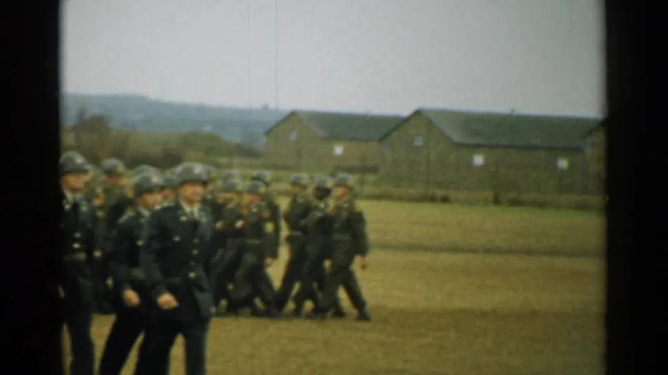 U.S. Army officers and soldiers march on a grassy field, with barracks and low hills visible in the background.