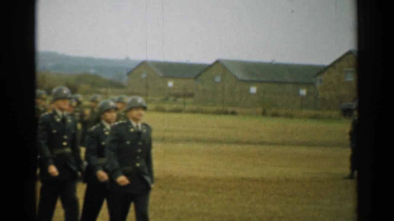Soldiers in dark uniforms and helmets march across a grassy field near barracks.
