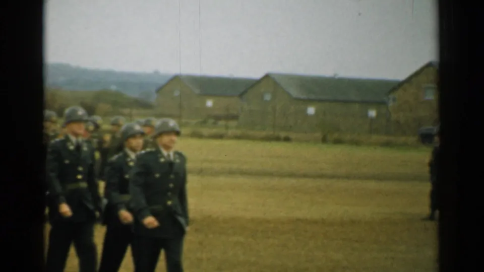 In New Hampshire 1952 footage, soldiers in dark uniforms and steel helmets march past brick barracks on a grassy base field.