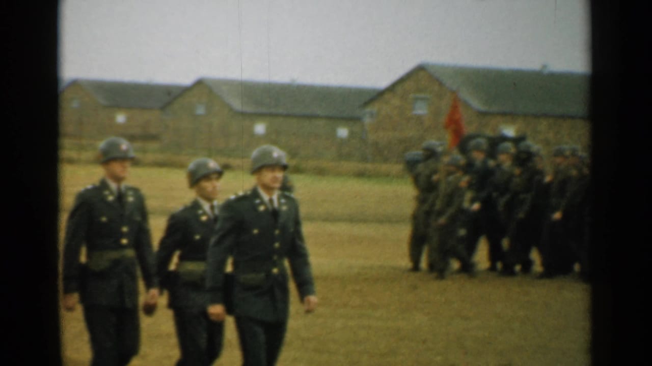 Three soldiers in helmets march across the base grounds while a larger uniformed group and a red flag appear in the background.