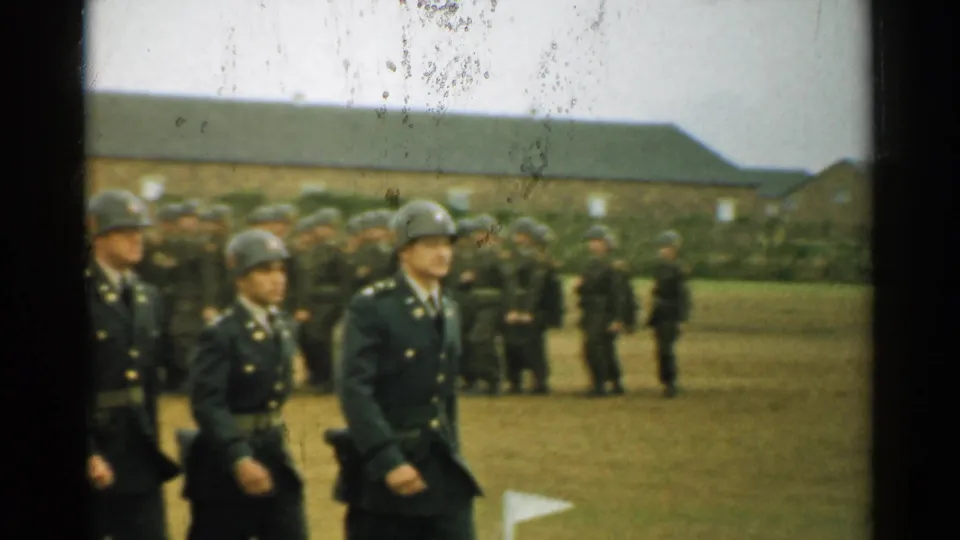 The formation continues across the grassy military field, with barracks, grouped soldiers, and visible 8mm film wear in the frame.