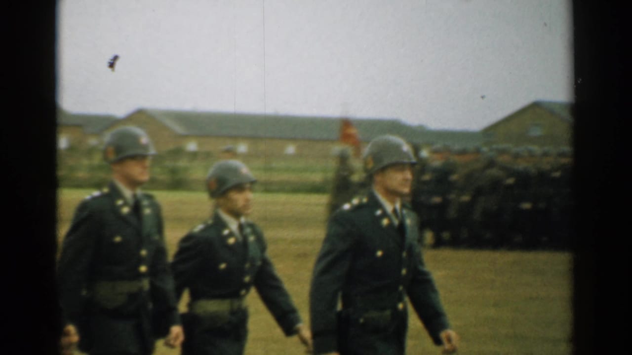 Three uniformed soldiers march past rows of troops and base buildings.