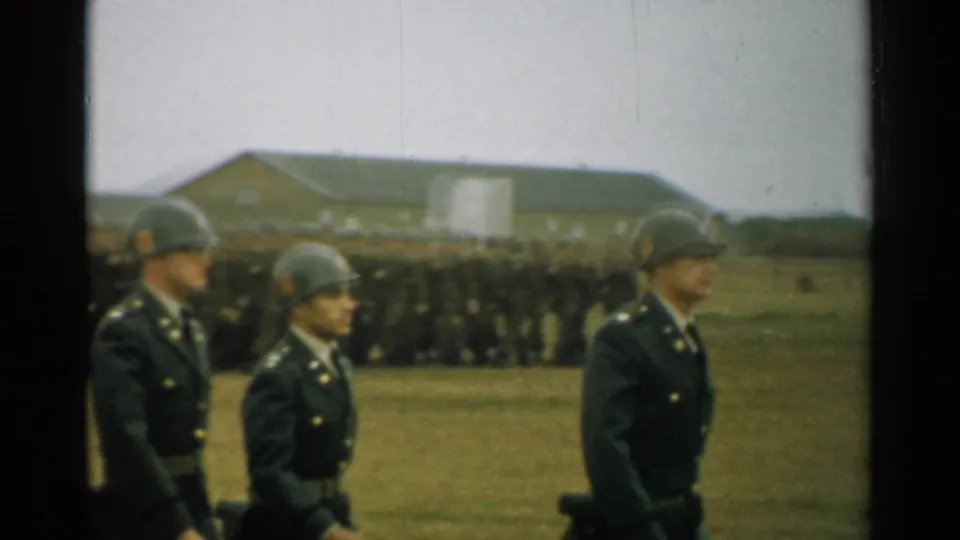 1952 New Hampshire 8mm frame showing soldiers marching near a larger formation, with long low buildings across the field.