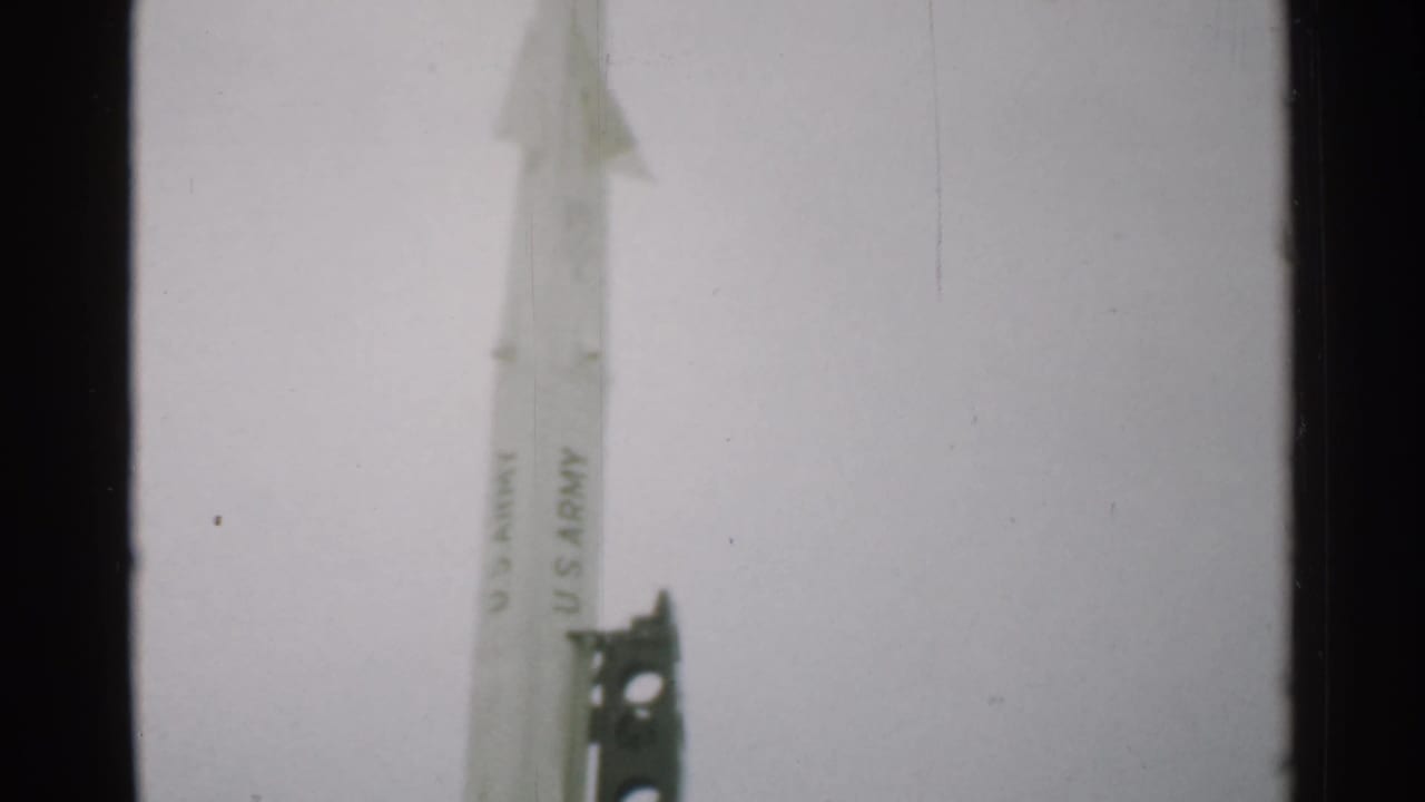 Upper section of a white U.S. Army-marked rocket beside a dark launch support against a pale sky.