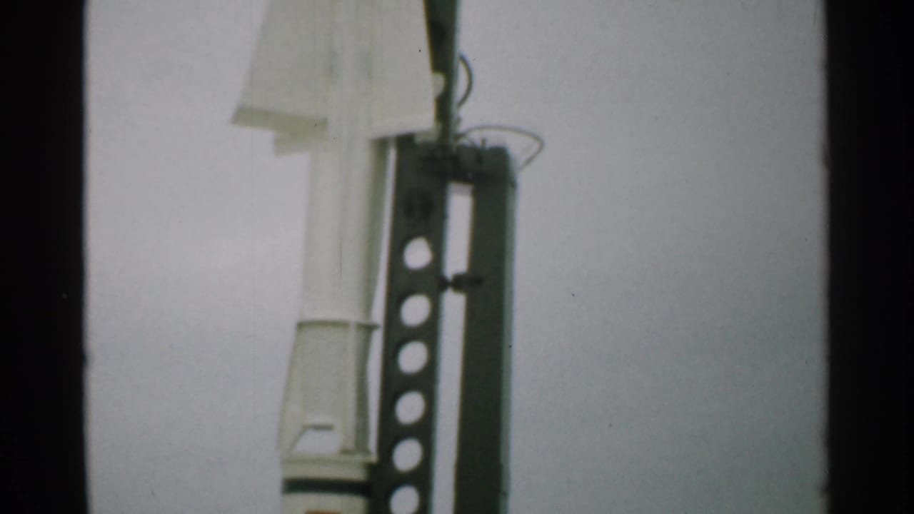 White rocket partly visible beside a dark metal launch frame against pale sky.