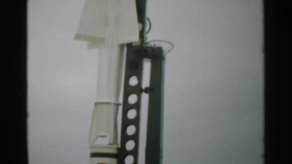 A 1952 New Hampshire 8mm frame shows the upper section of a white rocket beside a perforated metal launch frame outdoors.