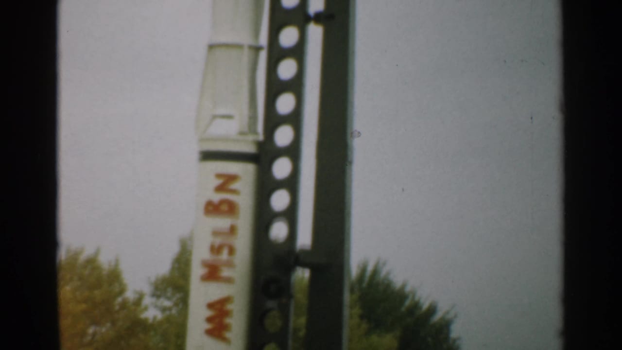 White rocket with red lettering stands beside a perforated metal launch frame.