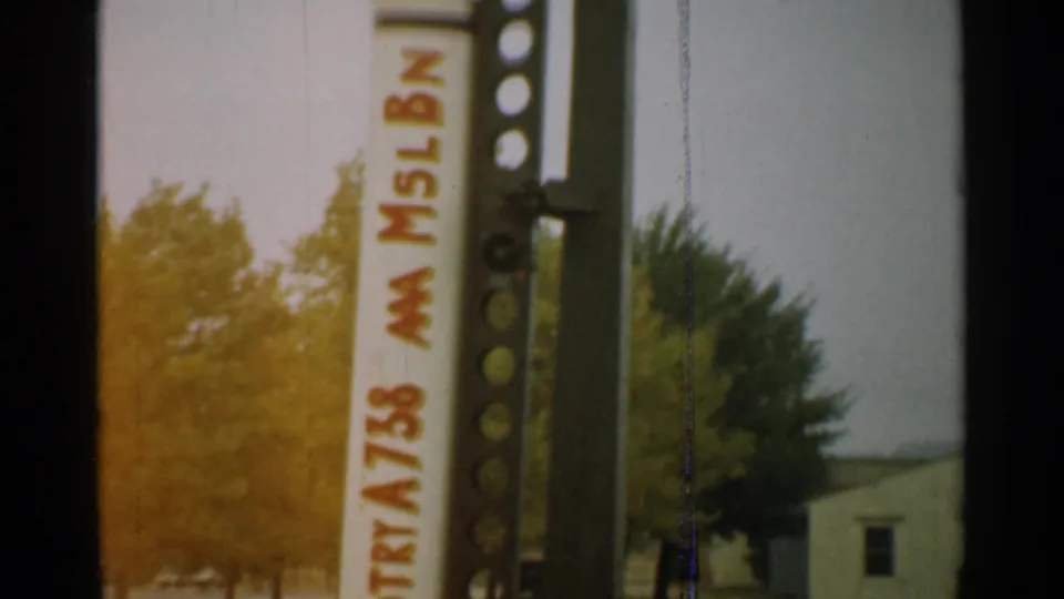 A lower angle shows the rocket body with red lettering, the metal stand, grass, trees, and a small background building.