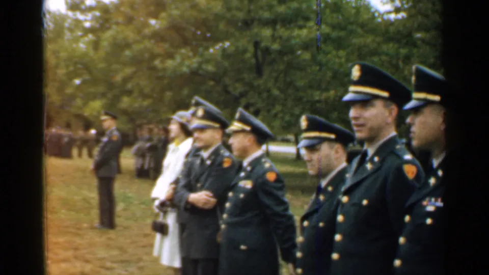 1952 New Hampshire home-movie frame of uniformed officers and a woman in a white coat standing in a grassy park.