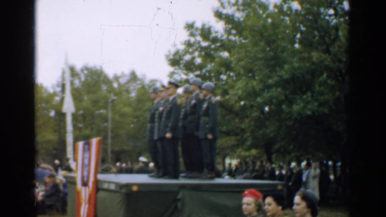 Uniformed men stand saluting on an outdoor platform while civilians watch below.