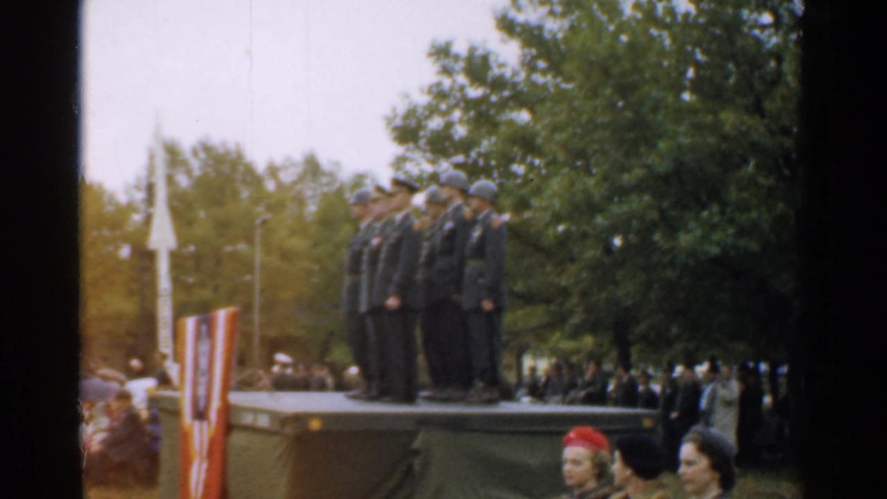 Uniformed men salute on an outdoor platform in New Hampshire, with spectators, trees, and a rocket-like display nearby.