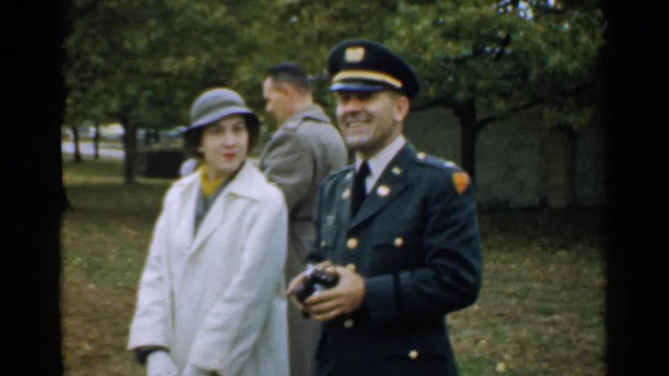 A uniformed man holding a camera smiles beside a woman in a light coat and hat during the New Hampshire event.