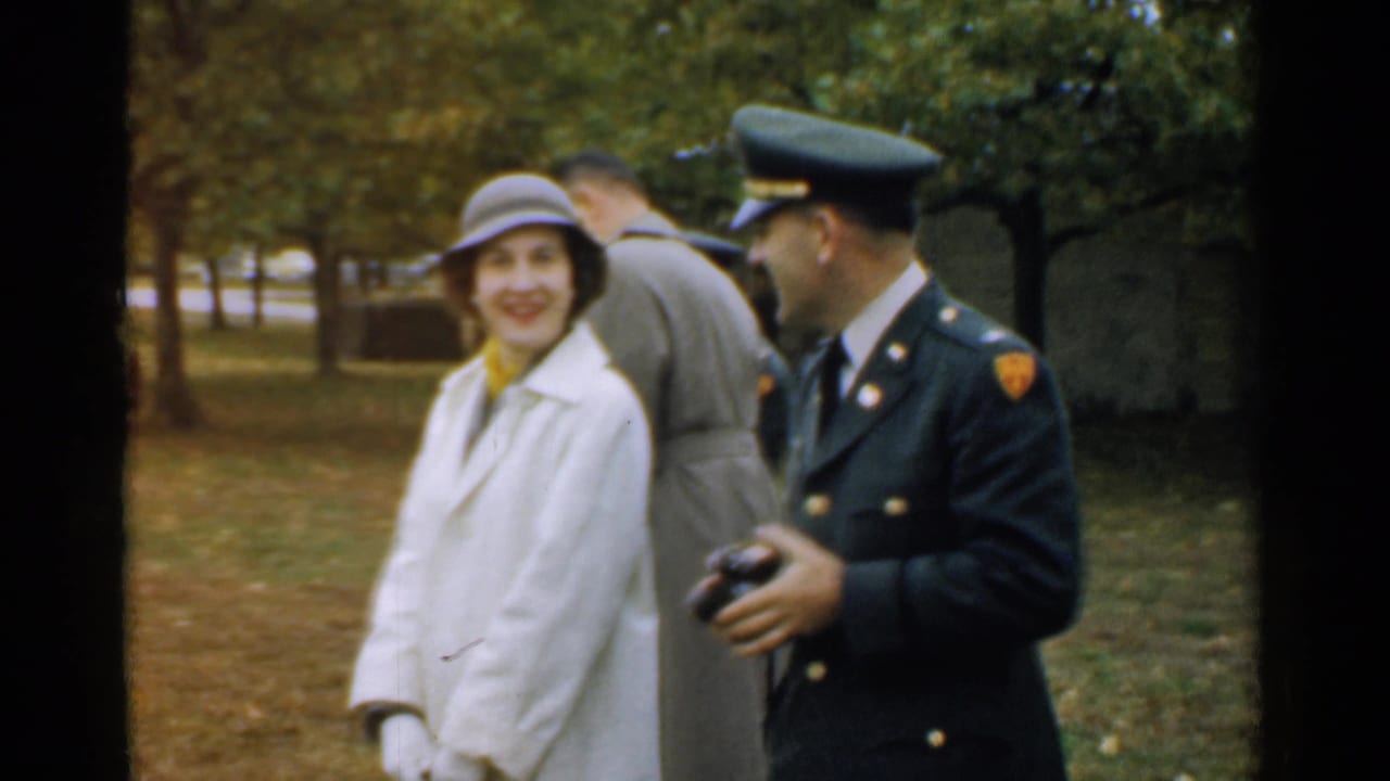 Woman in a light coat smiles beside a uniformed man holding a small camera in a park.