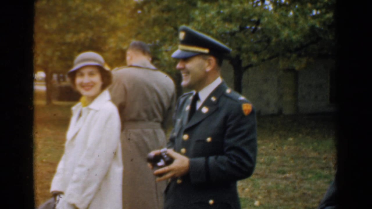 Smiling woman and uniformed man stand outdoors, with the man holding a camera.