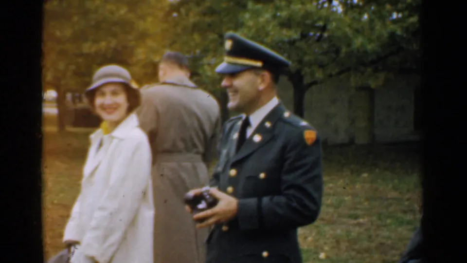 1952 New Hampshire frame of a laughing uniformed officer holding a camera beside a smiling woman in a light coat.