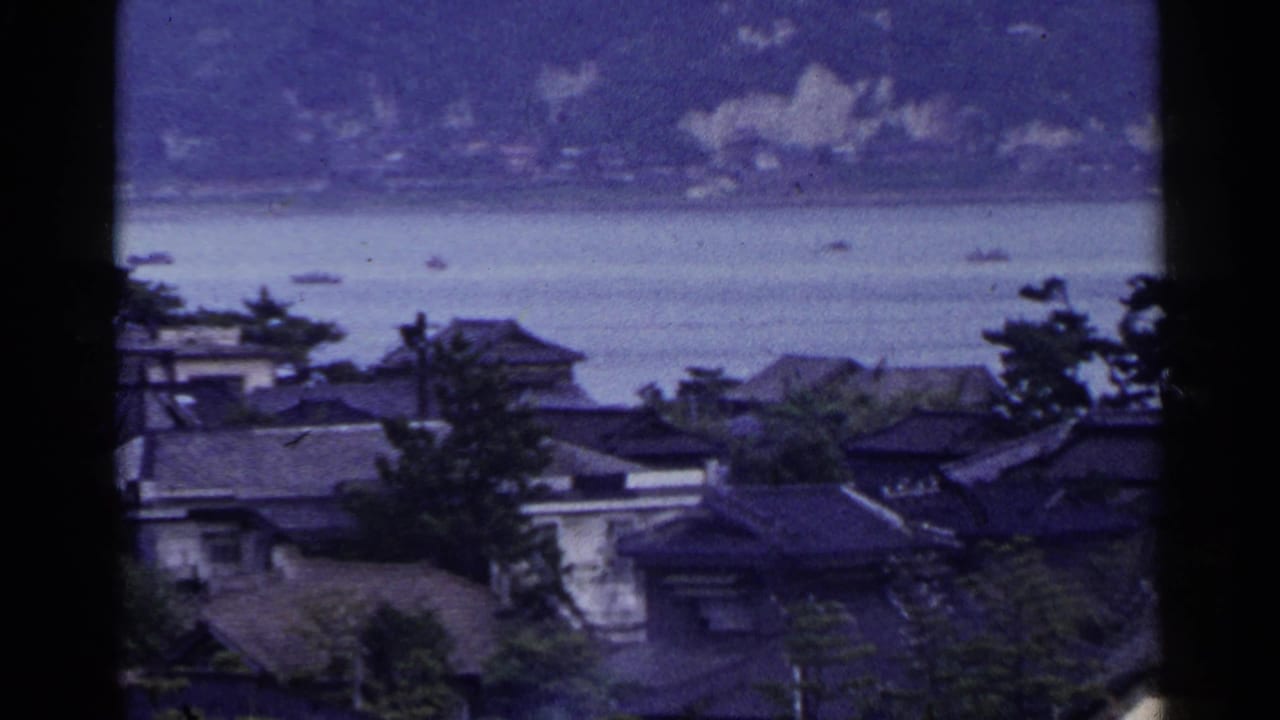 Coastal town roofs and trees overlooking calm water with small boats and distant hills.
