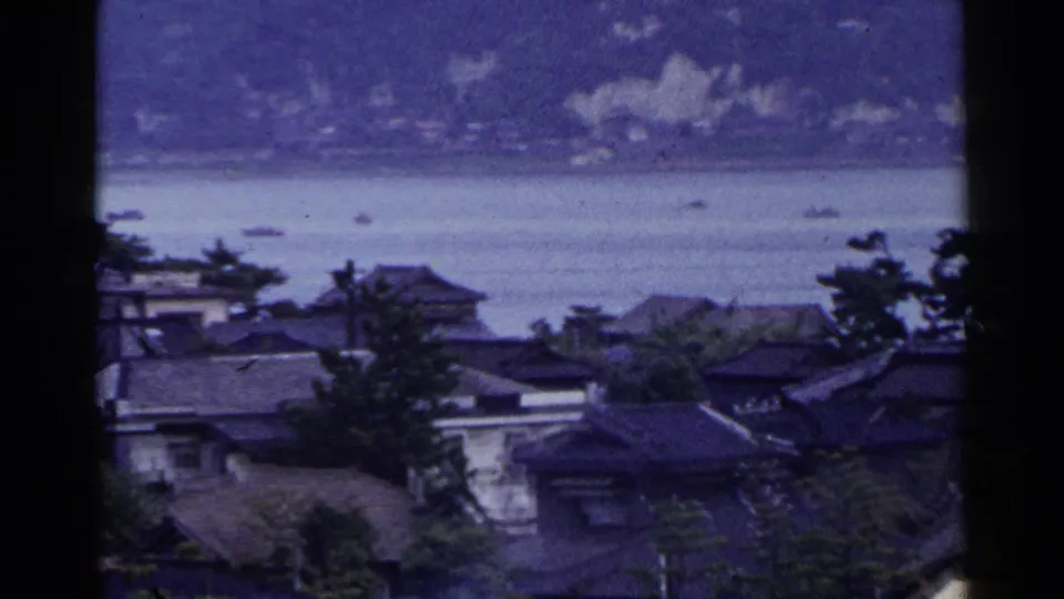 1953 Japan home-movie view of tiled-roof houses and trees beside calm coastal water, with small boats and hazy hills beyond.