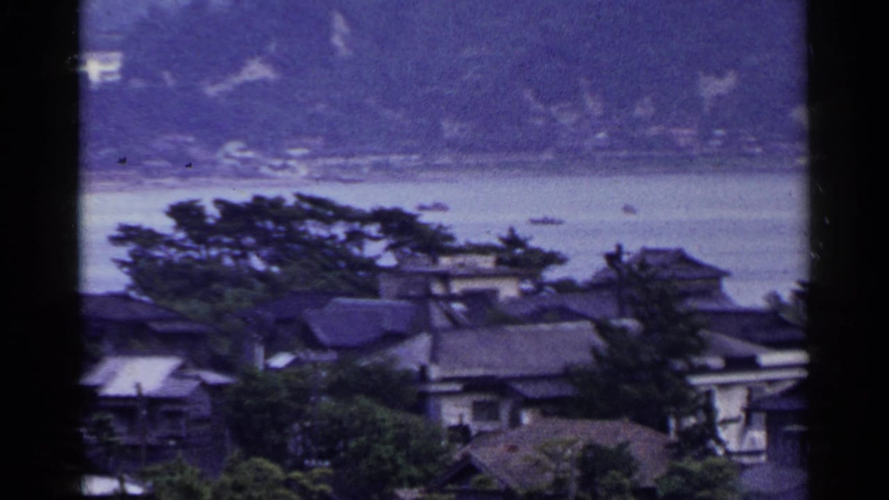 Waterfront houses and trees seen across calm water with boats and wooded hills in the distance.