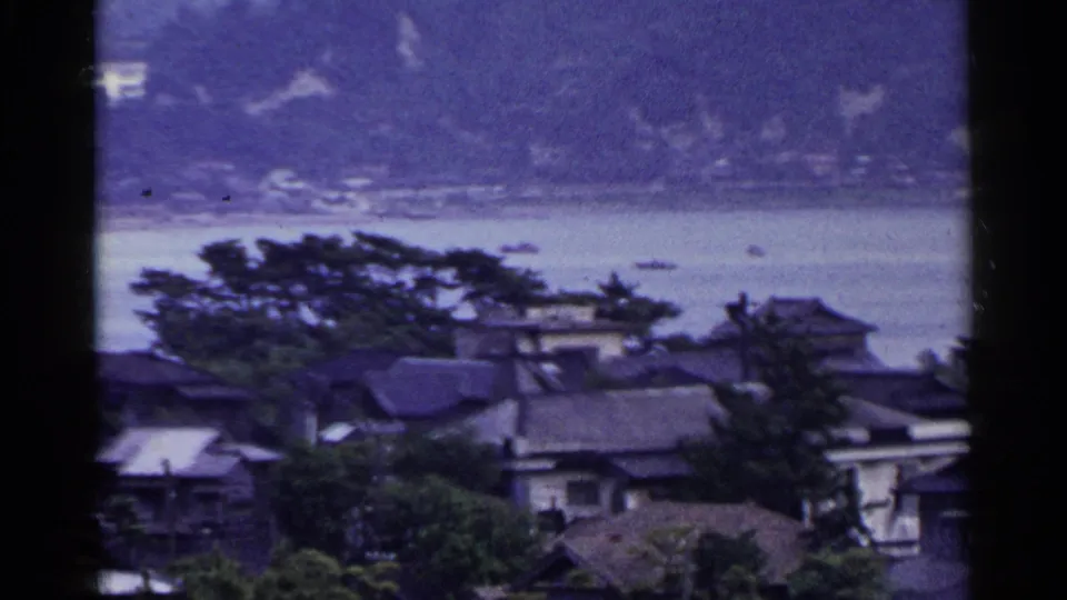 Elevated 1953 Japan view across waterfront homes, trees, small boats, and layered hills under a muted sky.