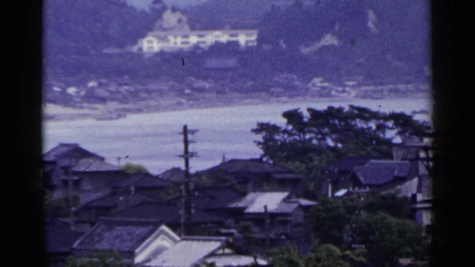 1953 Japan coastal view with rooftops, utility poles, trees, still water, and a pale hillside building in the distance.