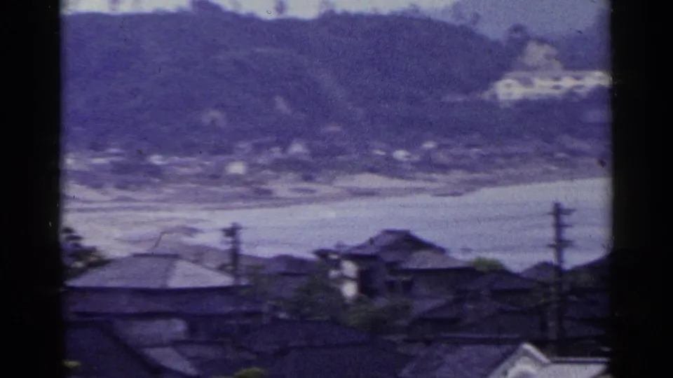 1953 Japan home-movie frame showing dark tiled roofs and utility poles below calm water and a broad wooded hill.