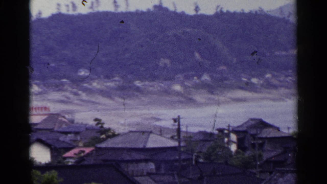 1953 Japan 8mm view of a hillside seaside town with rooftops, utility poles, trees, and a pale shoreline beyond.