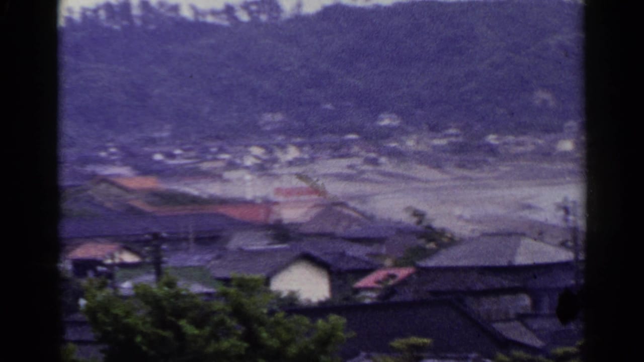 1953 Japan home-movie frame looking over rooftops toward a wooded hillside and pale inlet or beach area.