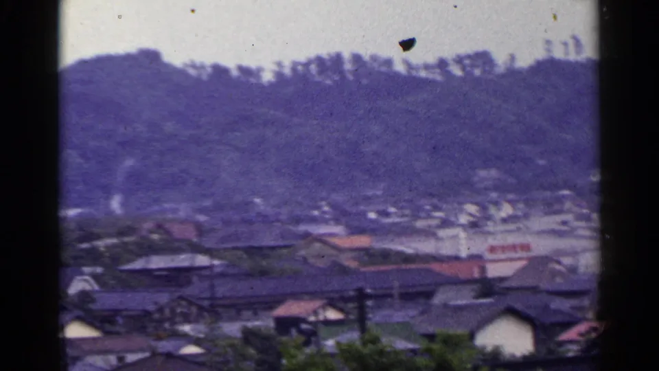 Soft 8mm view in Japan showing rooftops, a shoreline or inlet, and a hazy hillside beyond.