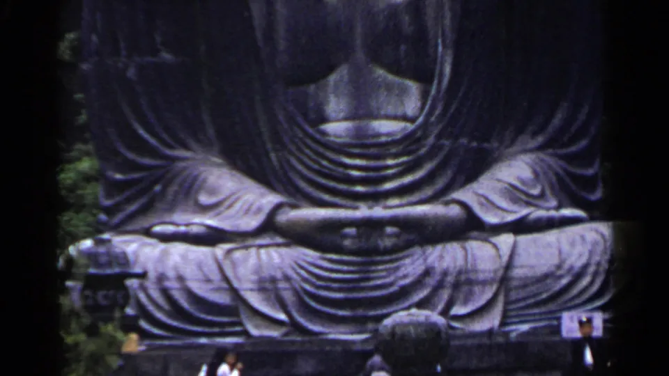 1953 Japan frame of a large seated metal Buddha statue, cropped at the torso, with trees and small visitors near the base.