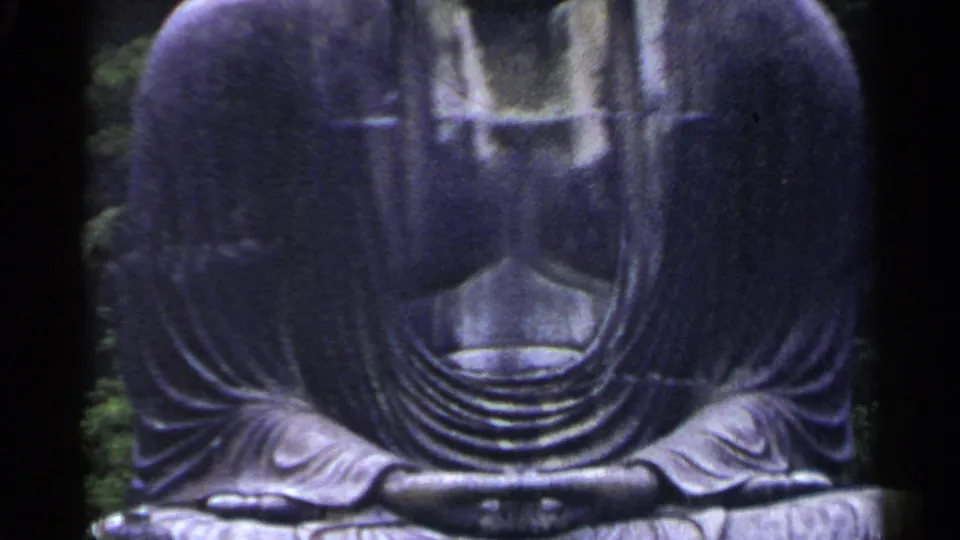 1953 Japan 8mm frame showing the lower torso and folded hands of a large dark gray outdoor Buddha statue.