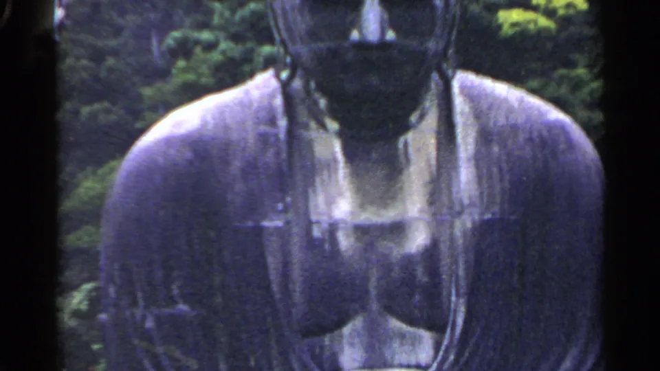 1953 Japan 8mm frame with the upper body of a large dark gray Buddha statue partly cropped beside green trees.