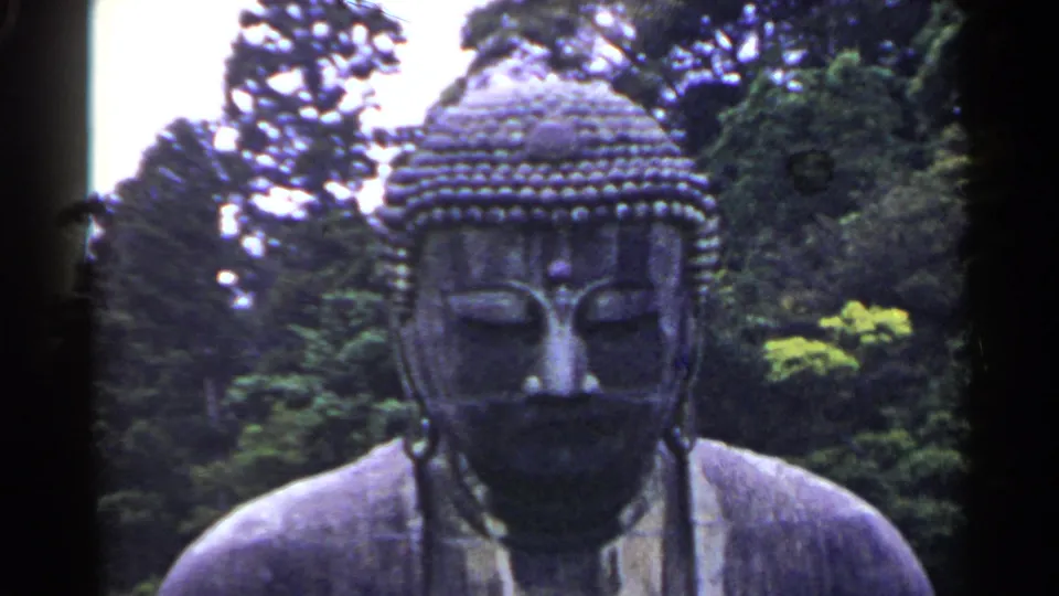 1953 Japan 8mm frame of a large dark gray Buddha statue outdoors, framed with trees and bright sky behind it.